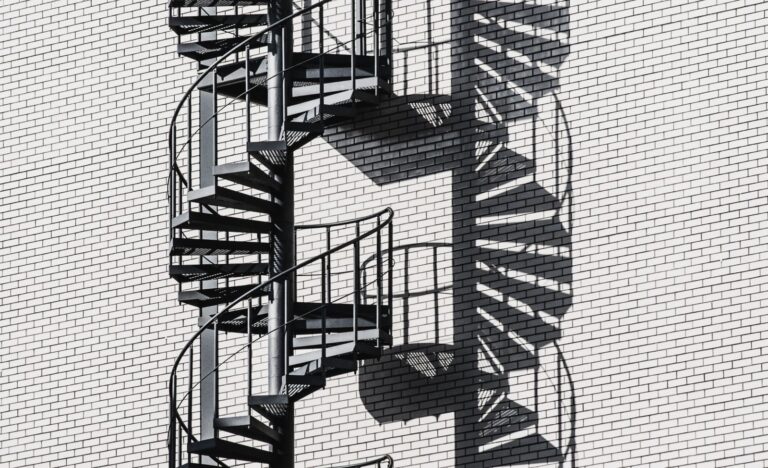 De esthetiek en duurzaamheid van metalen trappen en deuren in huis Black spiral staircase casting intricate shadows on a white brick wall in bright daylight.