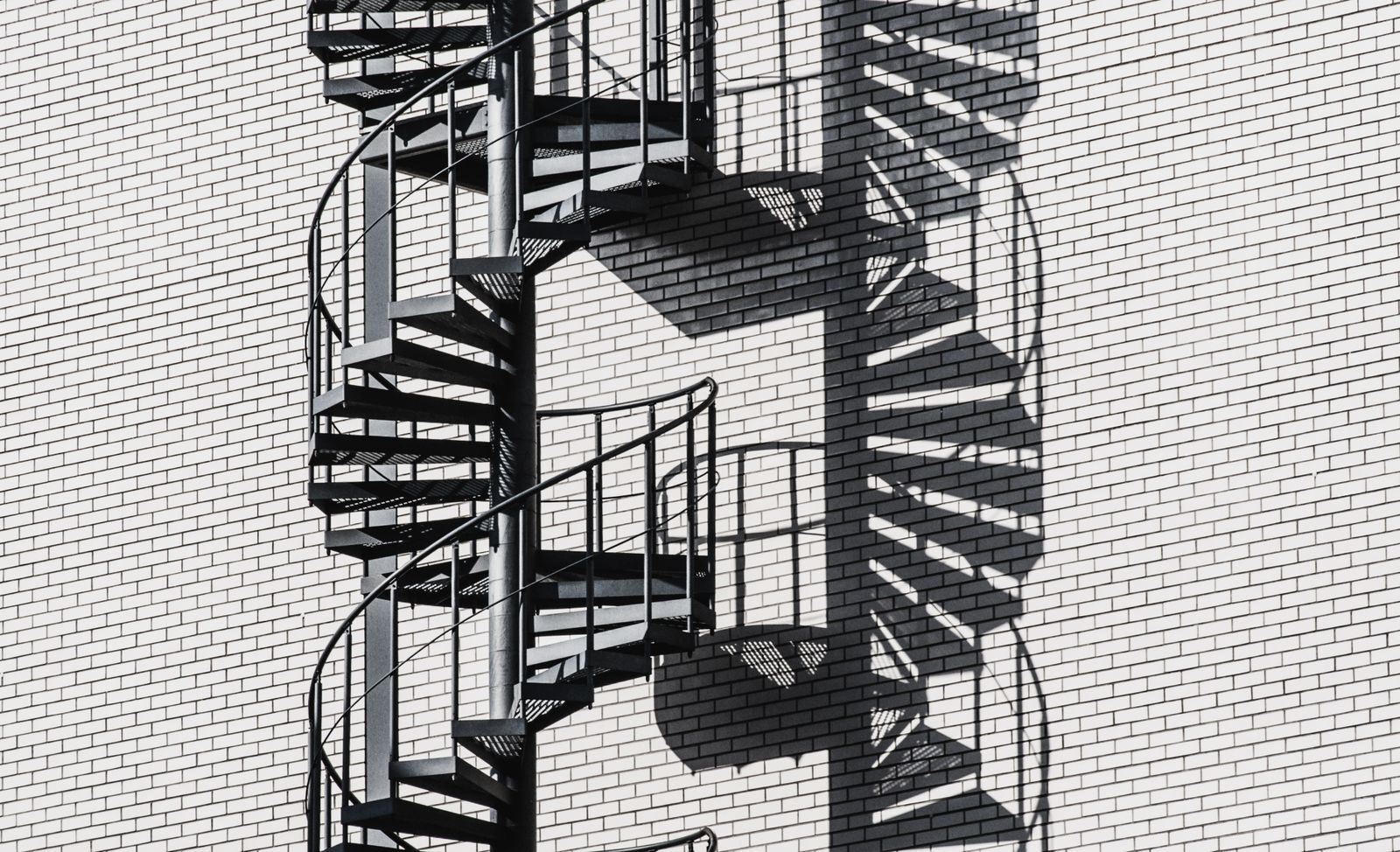 Black spiral staircase casting intricate shadows on a white brick wall in bright daylight.